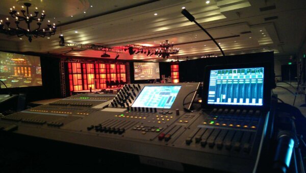 A view from the AV control booth at a corporate event, showing a digital audio mixing console with illuminated faders, knobs, and touchscreen displays for audio levels and mixing. In the background, a stage with a red and orange illuminated backdrop is framed by a truss structure supporting stage lights. Two large projection screens display an image of a banquet-style event setup. Chandeliers hang from the ceiling, and the room is arranged with seating for attendees. Overhead trusses support lighting and audio equipment for the event.