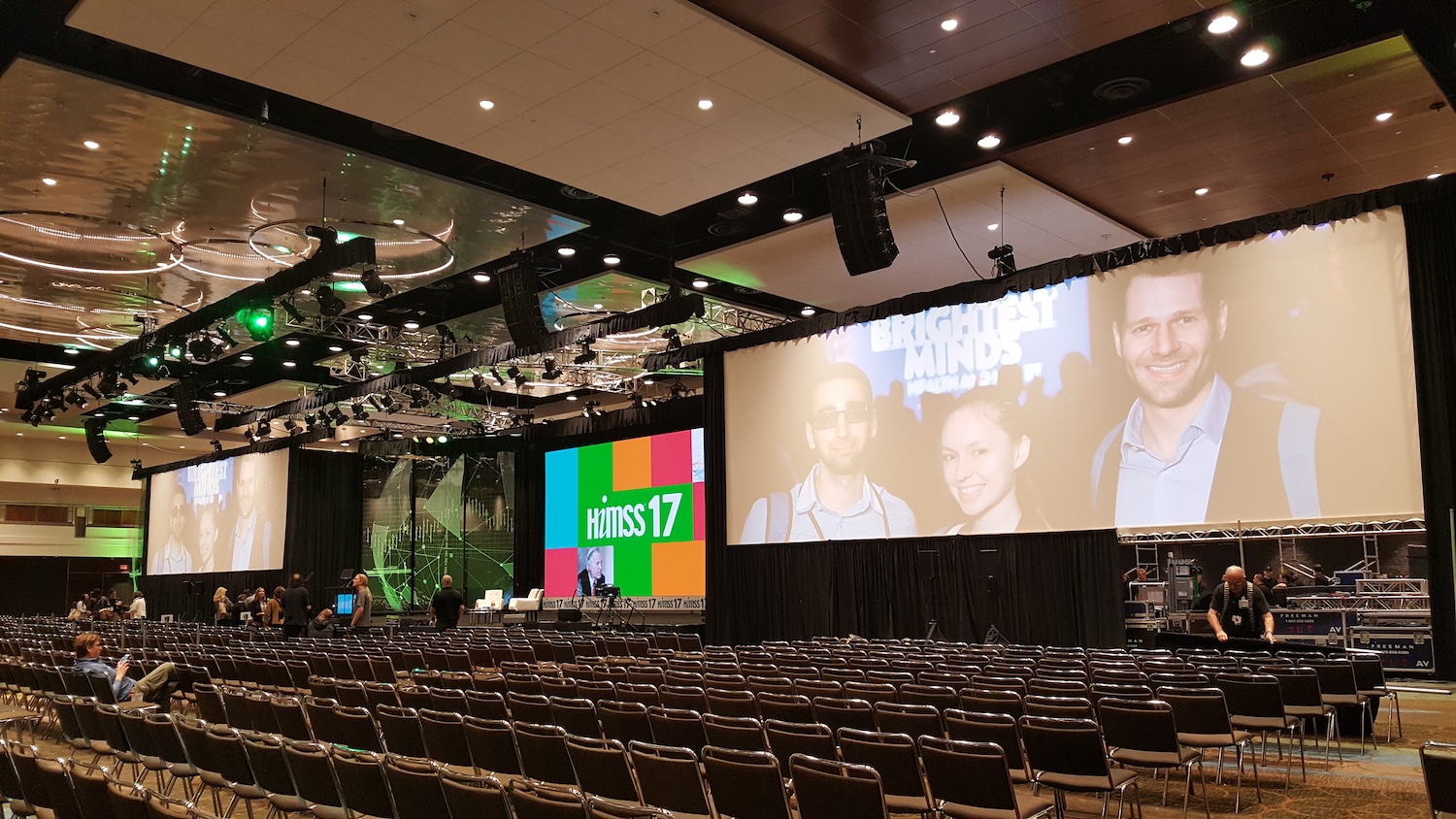 A conference hall set up with chairs and a stage with large screens, one displaying 'himss 17' and 'Brightest Minds' graphics, with lighting rigs overhead.
