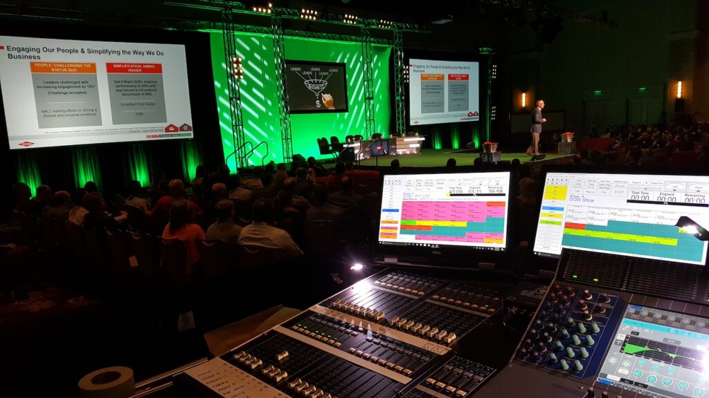 A view from a sound engineer's booth at a conference, with a speaker on stage and presentation screens visible to the audience.