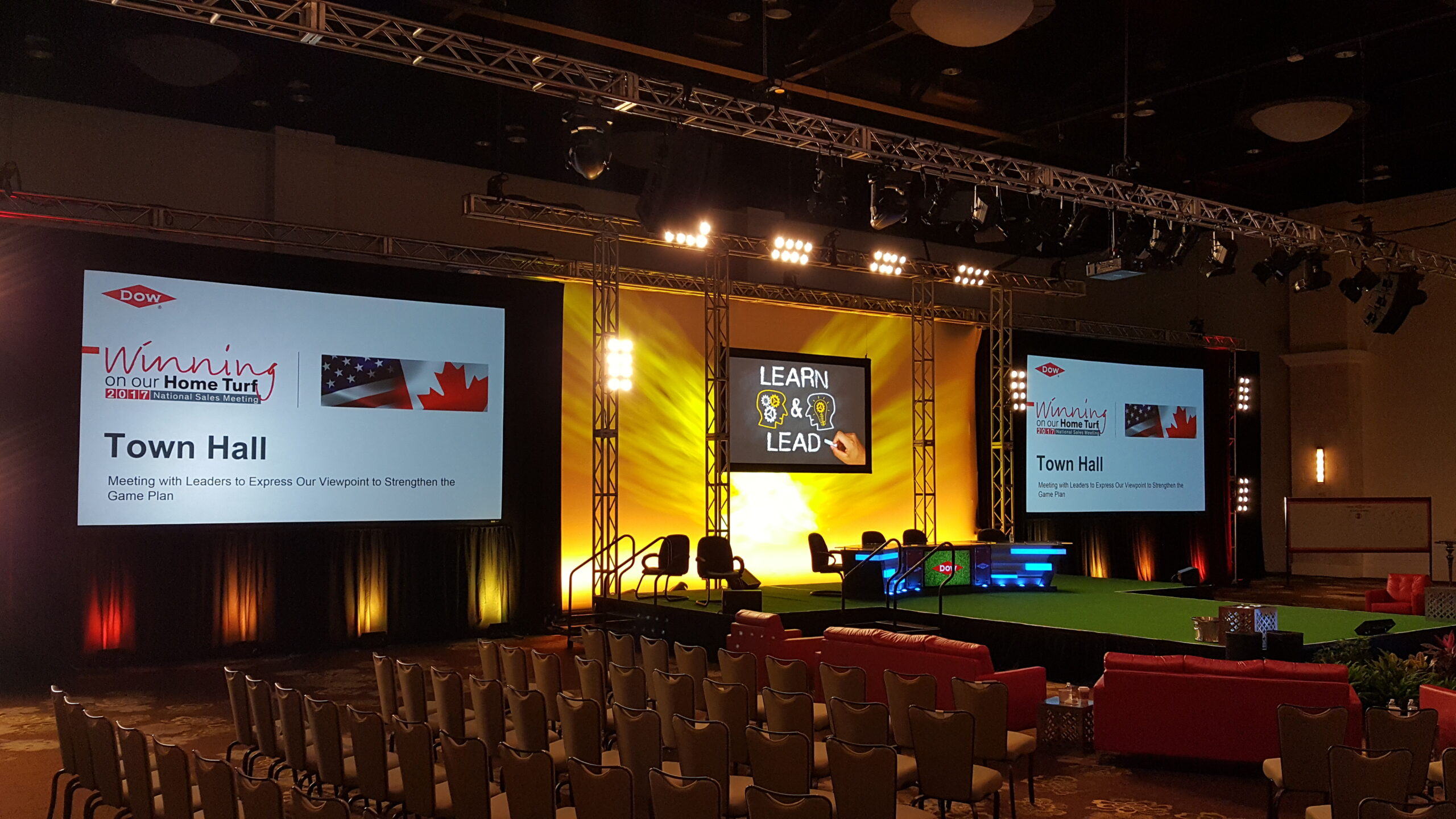 A stage set up for a town hall meeting with large screens displaying "Winning on our Home Turf" and "Learn & Lead" in a room filled with rows of chairs.