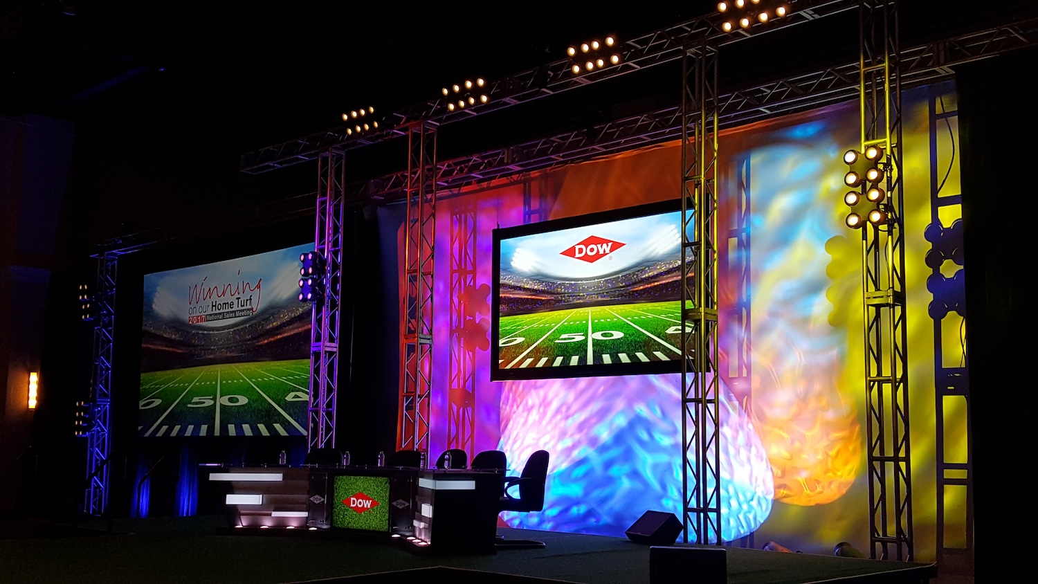 An event stage with colorful lighting, featuring screens with 'Winning on our Home Turf' and the Dow logo, set for a presentation or conference.
