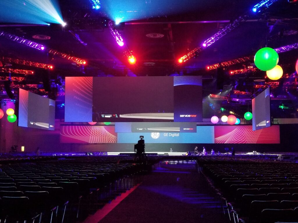 An event space with rows of empty chairs facing a large stage with an intricate screen setup, lit by colorful lights and stage effects.