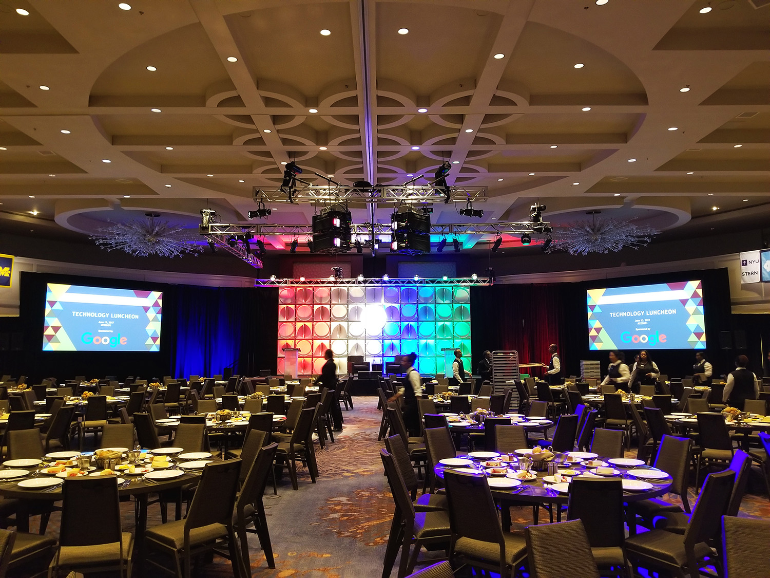 A banquet hall set for a 'Technology Luncheon' with Google branding on screens, arranged tables, and stage lighting equipment overhead.