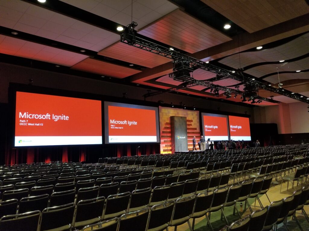 A large conference hall with rows of chairs and multiple screens displaying 'Microsoft Ignite' branding, set up for an event.