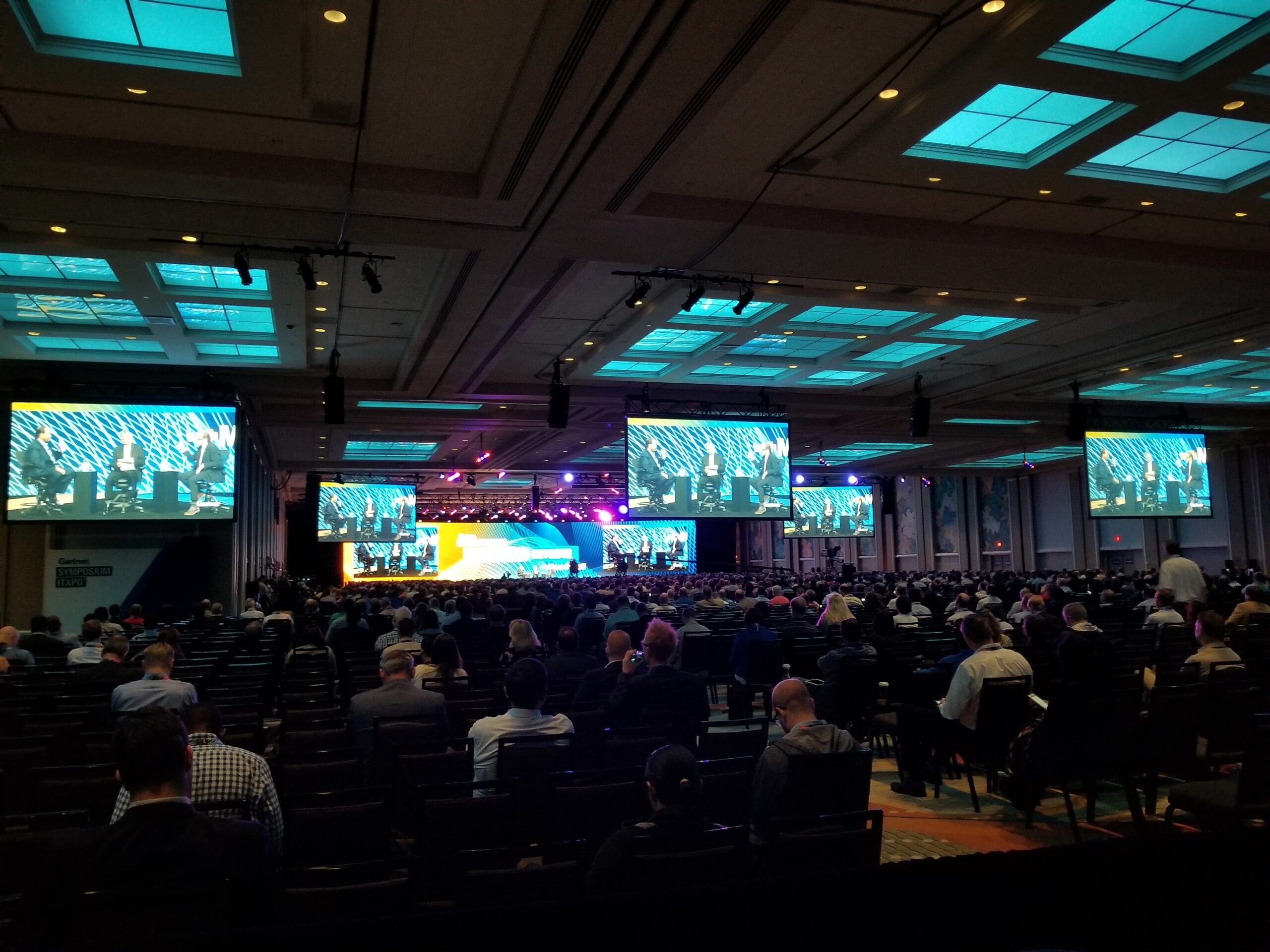 A conference event with a large audience, multiple screens showing a speaker, and a well-lit stage setting.