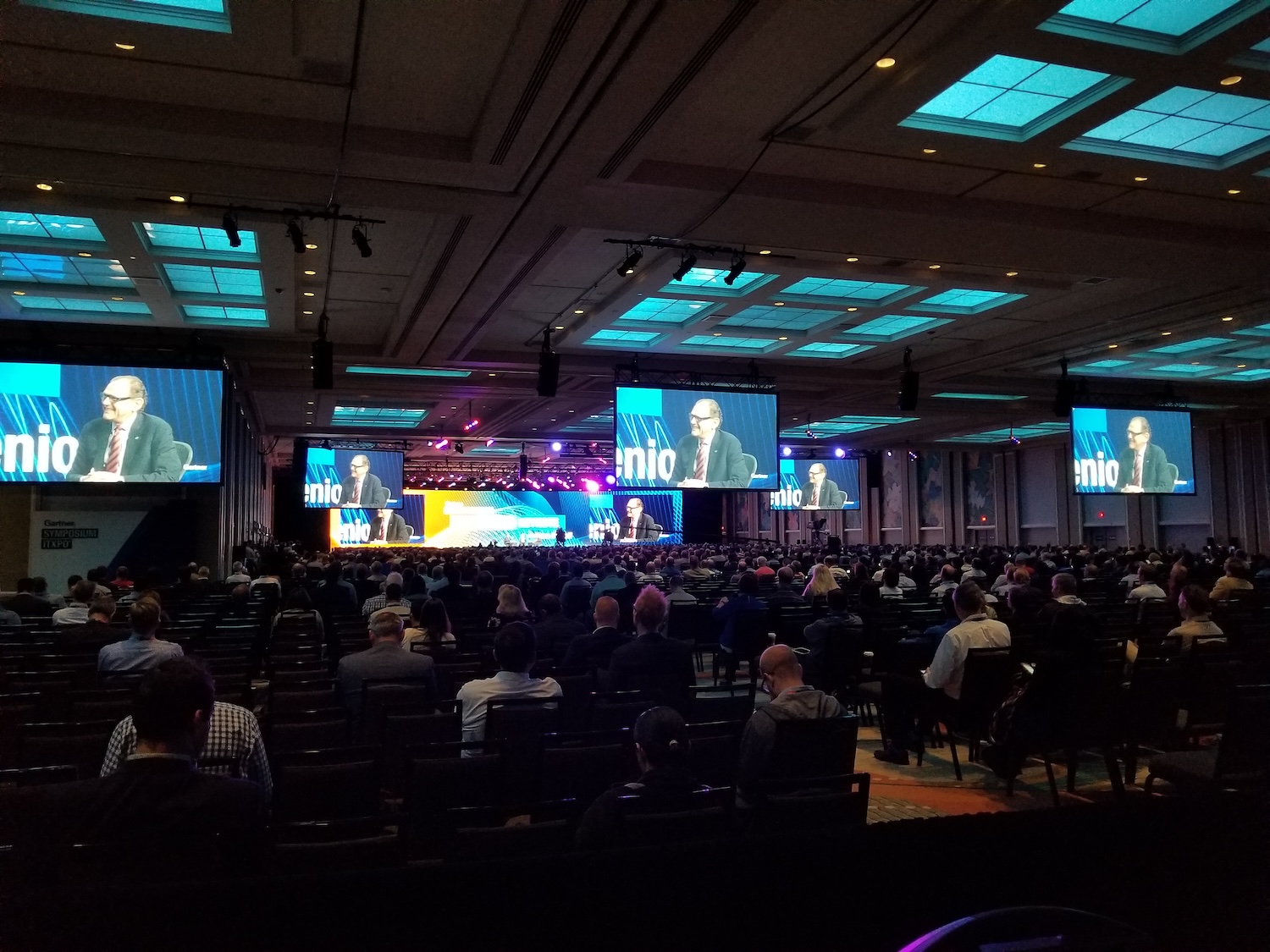 A conference event with a large audience, multiple screens showing a speaker, and a well-lit stage setting.