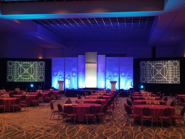 A corporate event stage with two podiums and a central backdrop lit with blue uplighting. Two large projection screens on either side display test patterns with geometric grid designs. The floor is covered in patterned carpet, and round tables with red tablecloths are set up for attendees. The AV setup includes stage lighting, projection screens, and sound equipment.