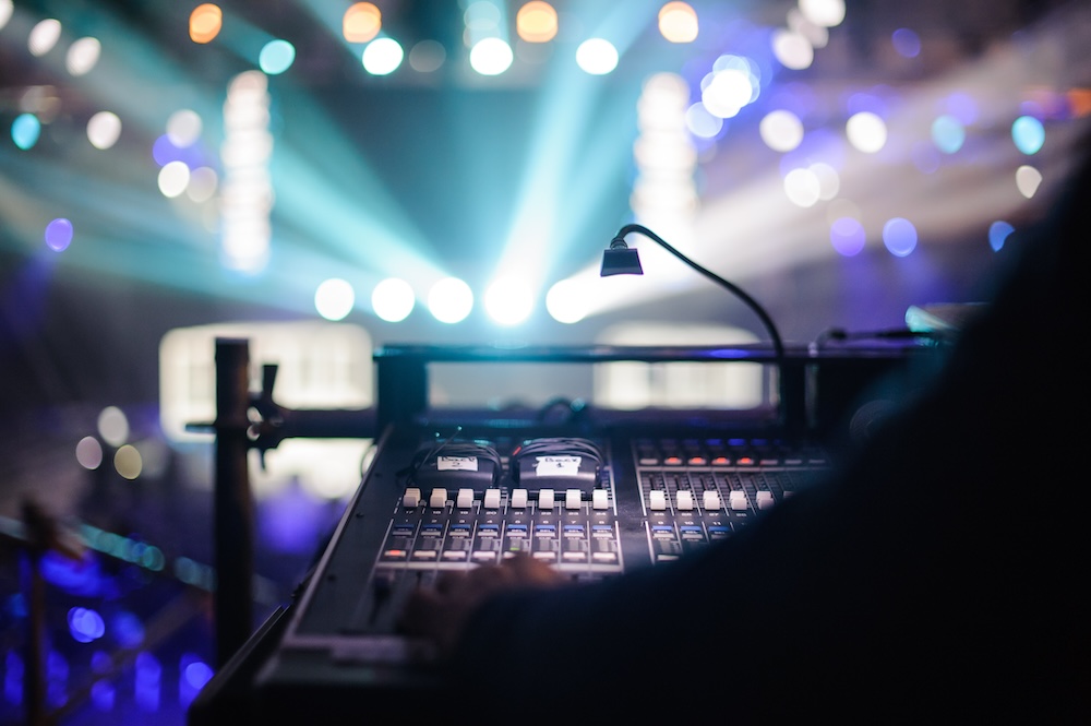 Close-up of a person's hand adjusting sliders on a sound mixer at a concert with vibrant stage lights in the background.