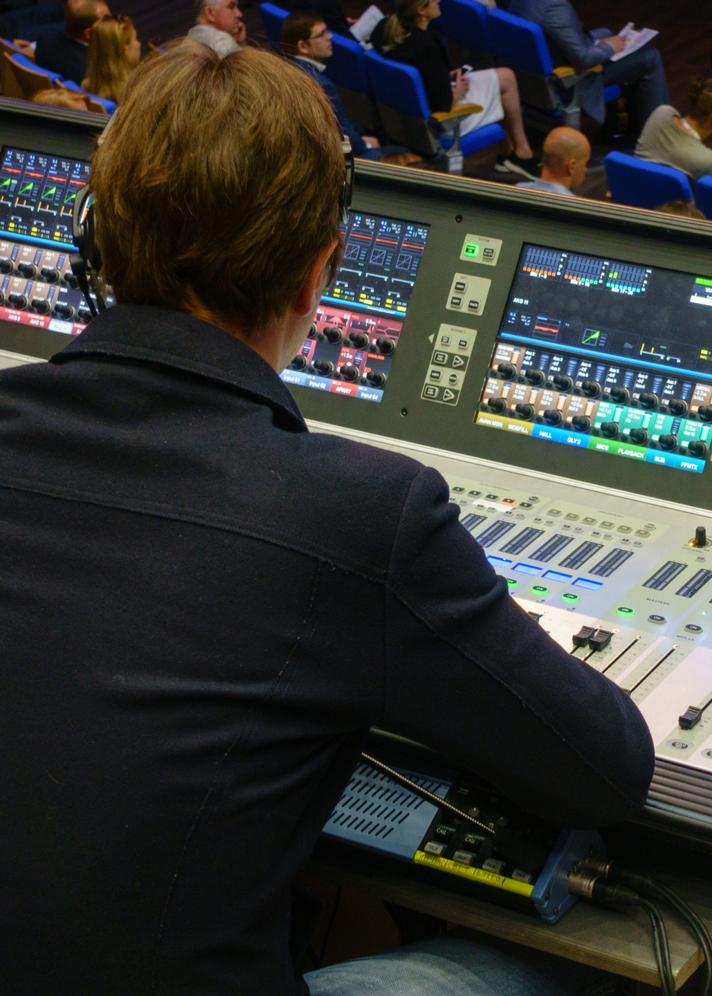 A V1 High-End Switcher from behind operating a large mixing console in a room with an audience in the background.