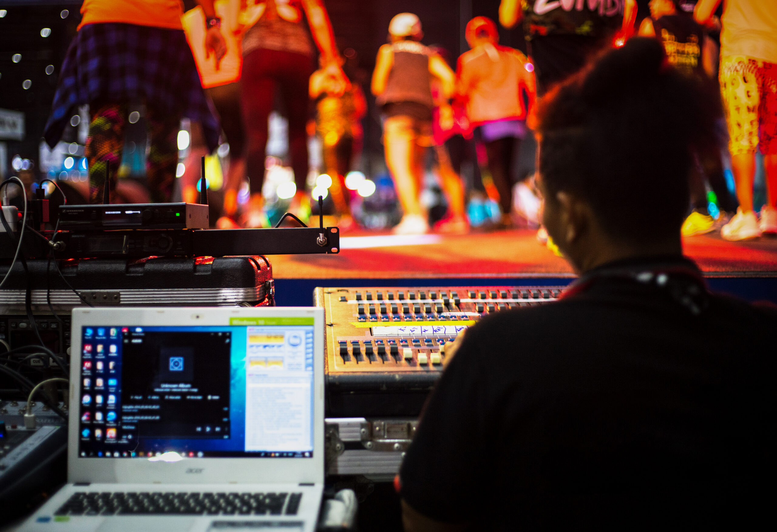 View from behind a sound technician at a mixing board with a laptop in the foreground and colorful stage lights illuminating performers in the background.