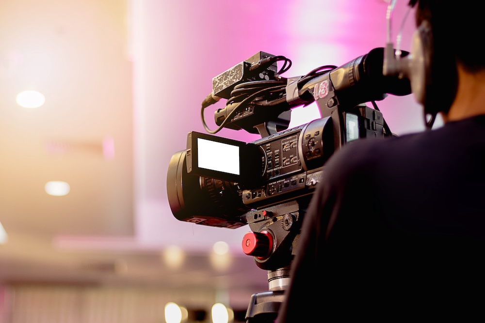 A professional video camera operated by a cameraperson, focused on a pink-lit stage or presentation area.