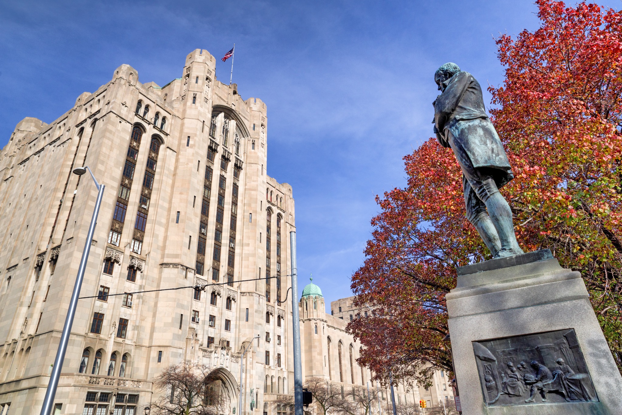 A statue of a historical figure stands prominently in front of an impressive art deco building under a clear blue sky. The building is adorned with tall windows and a U.S. flag at the top, while autumnal trees with red leaves frame the scene.