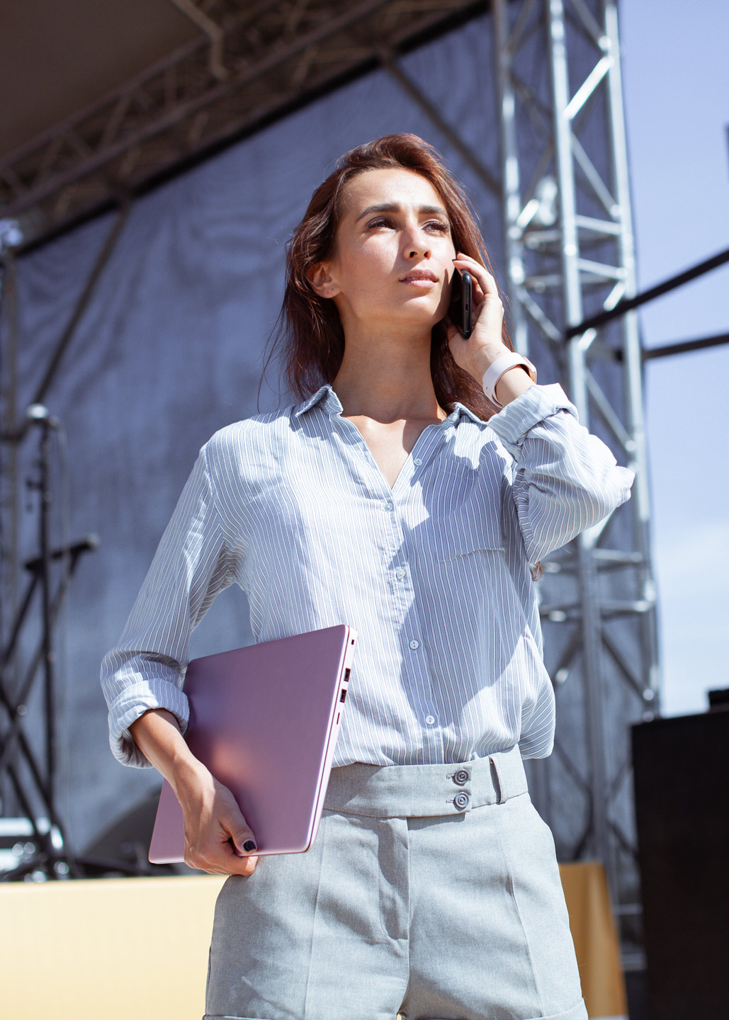 A woman on set of an outdoor event on the phone and holding her laptop.