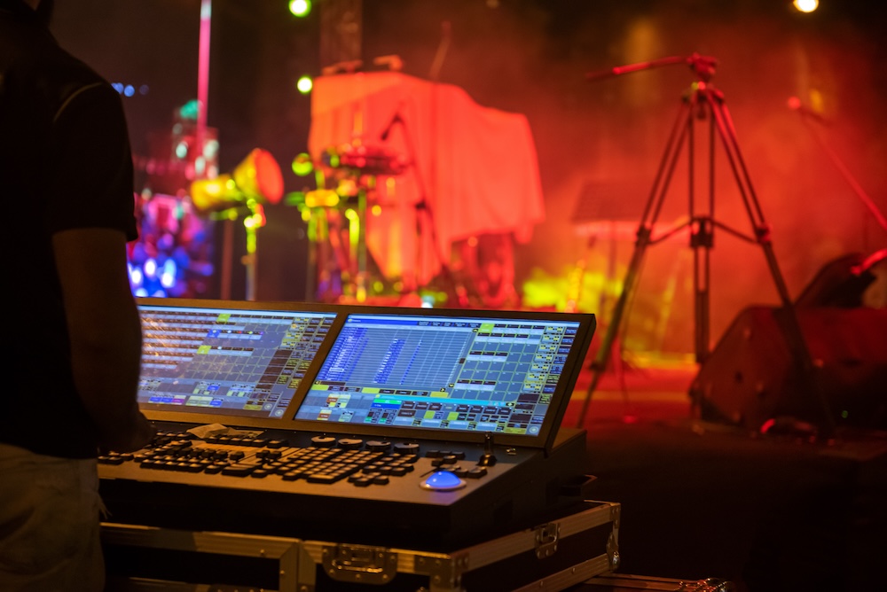 A sound engineer's digital mixing console at a live concert venue with colorful stage lights in the background.