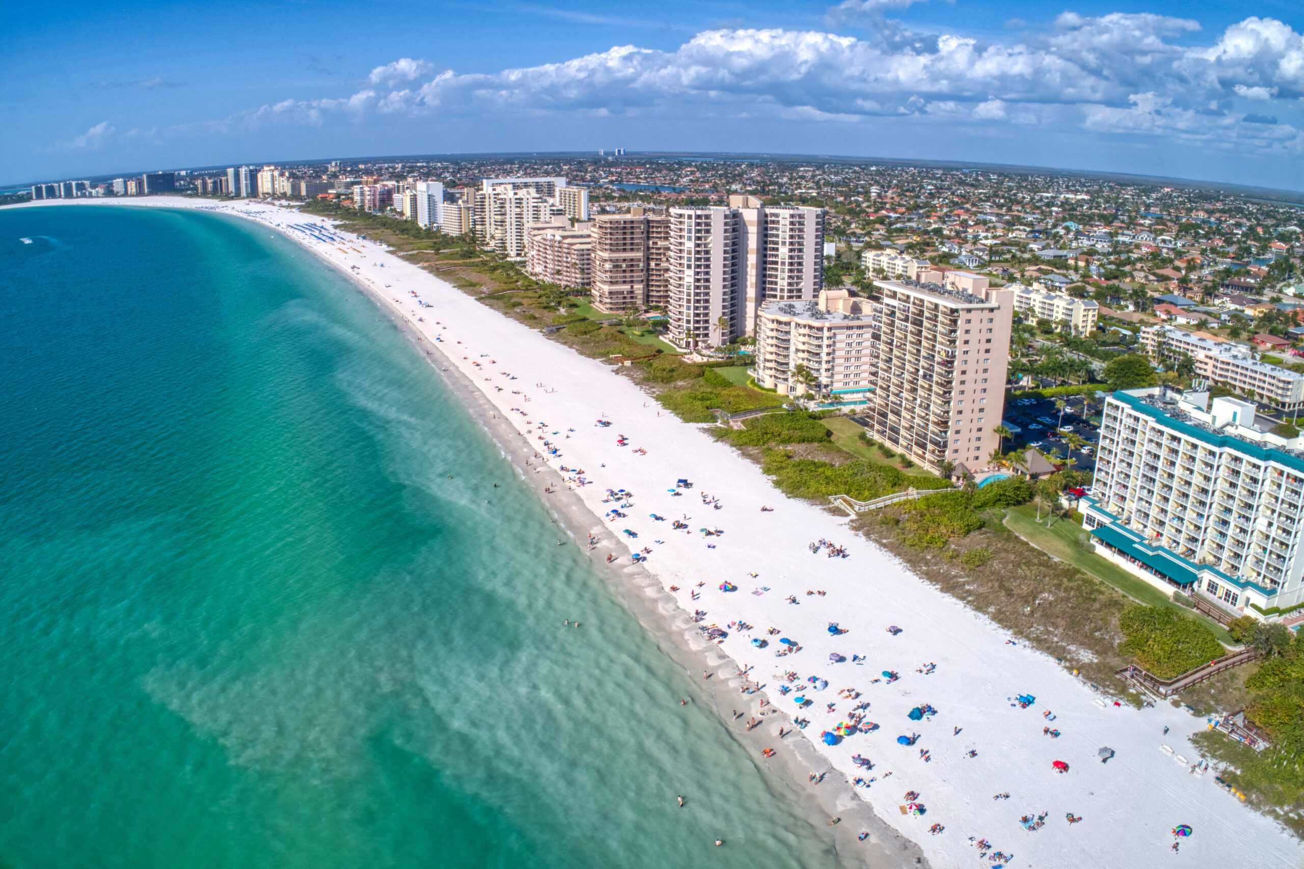 Aerial view of Marco Island, FL, showcasing a long stretch of white sandy beach lined with beachfront high-rise buildings. The clear turquoise water of the Gulf of Mexico contrasts with the bright, sunlit beach, where people are relaxing under colorful umbrellas.