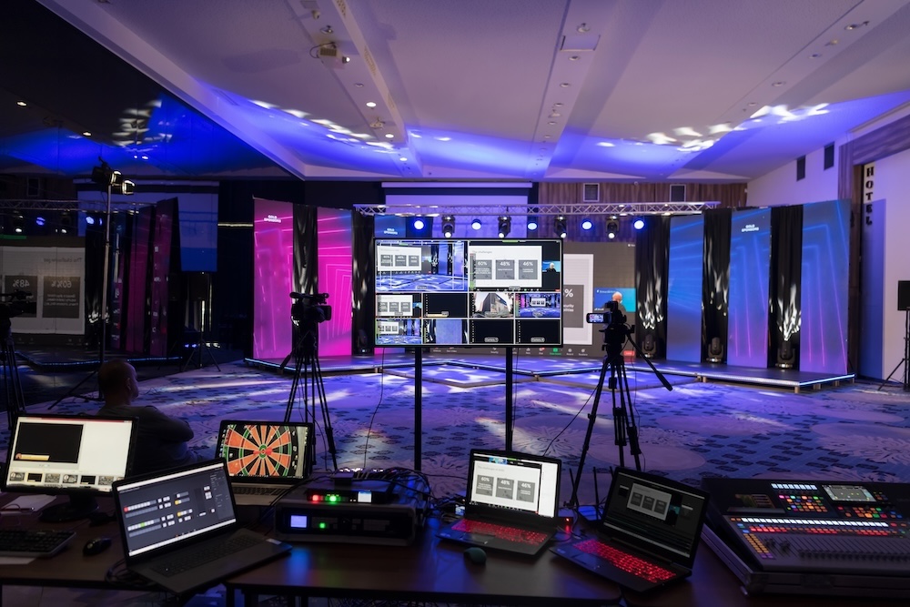 A live event control room setup with multiple monitors, laptops, a video camera on a tripod, and a stage with dynamic lighting in the background.