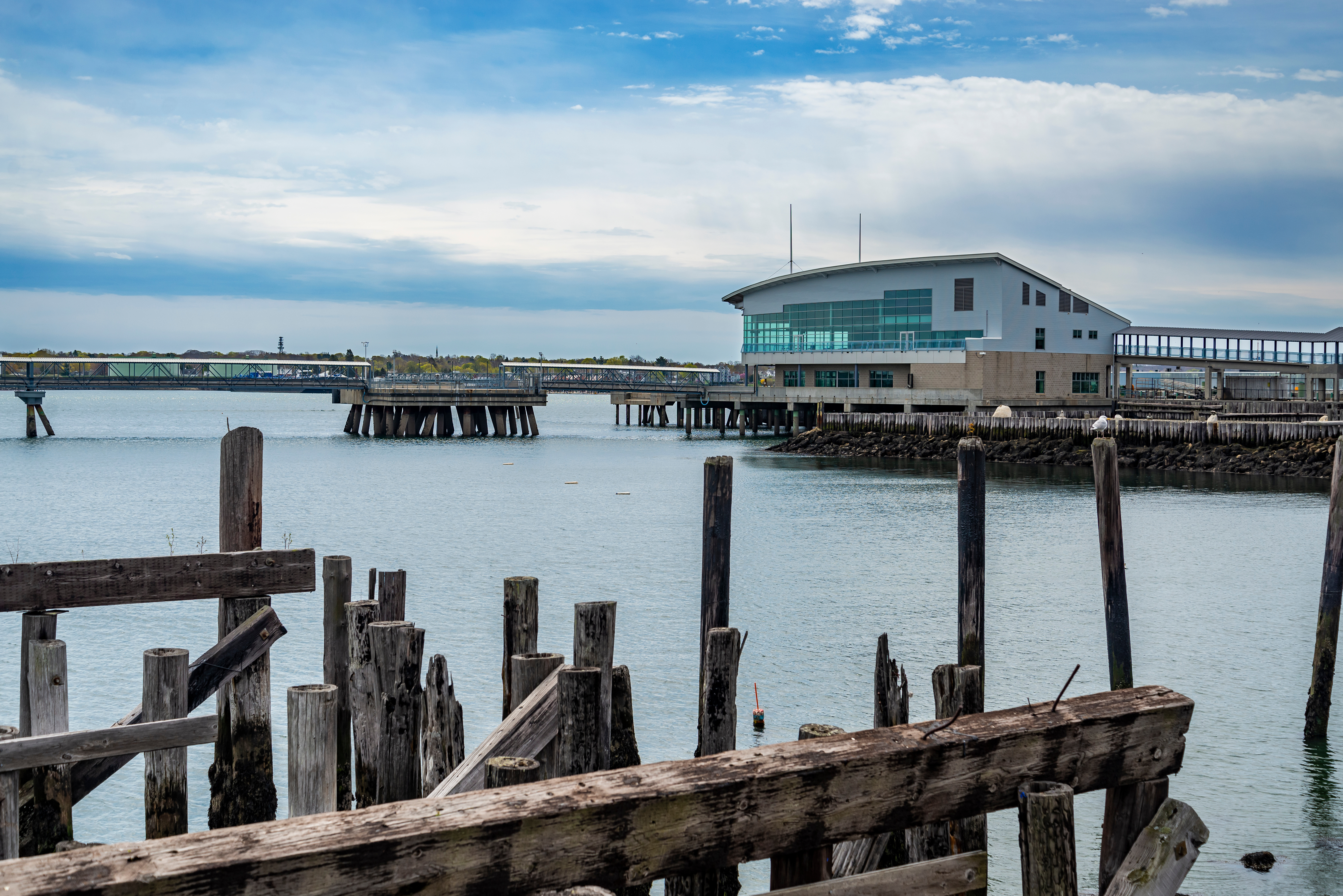 A modern building with large windows stands on a pier over calm water, with an old wooden structure in the foreground under a cloudy sky.