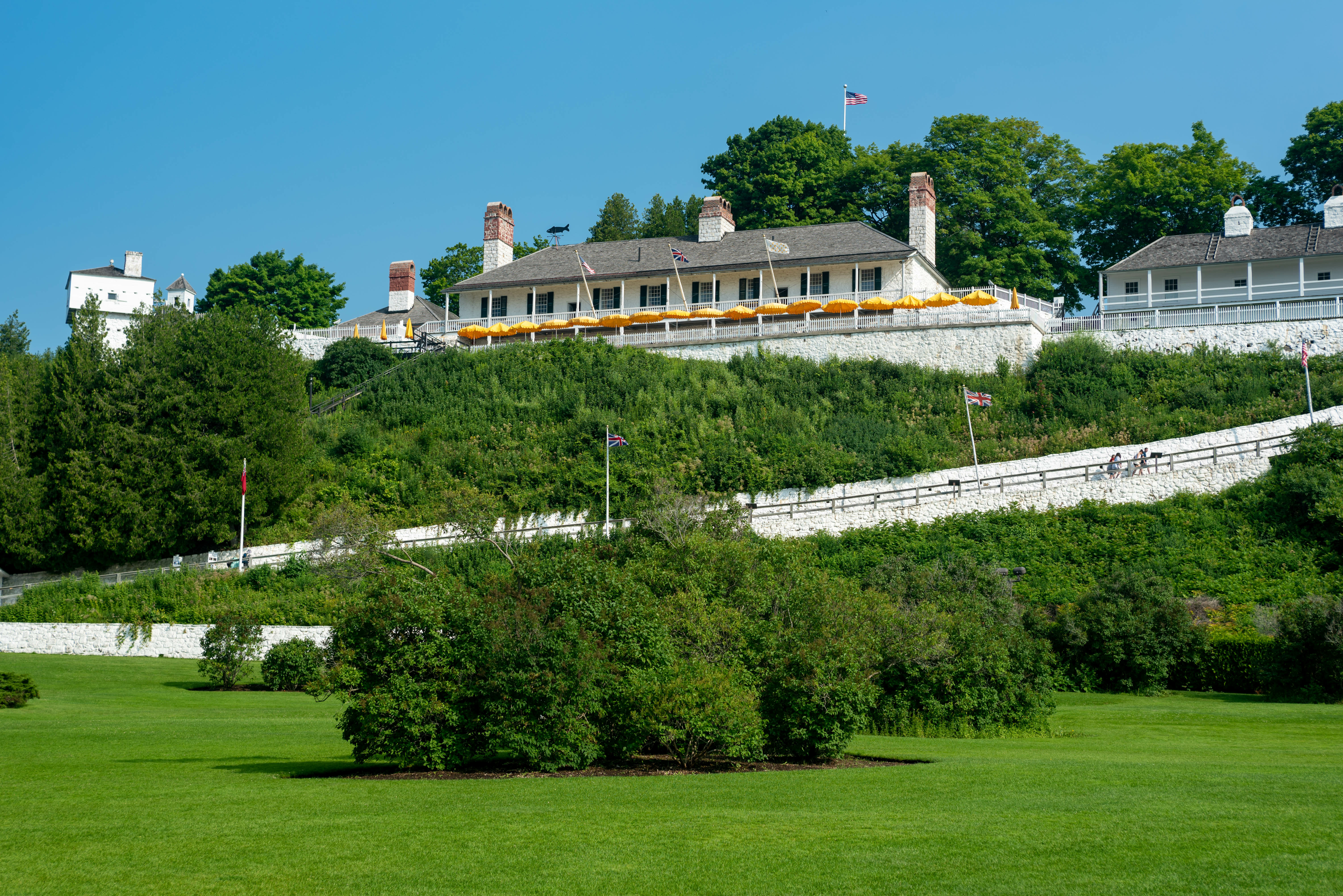 A historic fort on Mackinac Island, Michigan, features white buildings with red brick chimneys atop a lush green hillside.