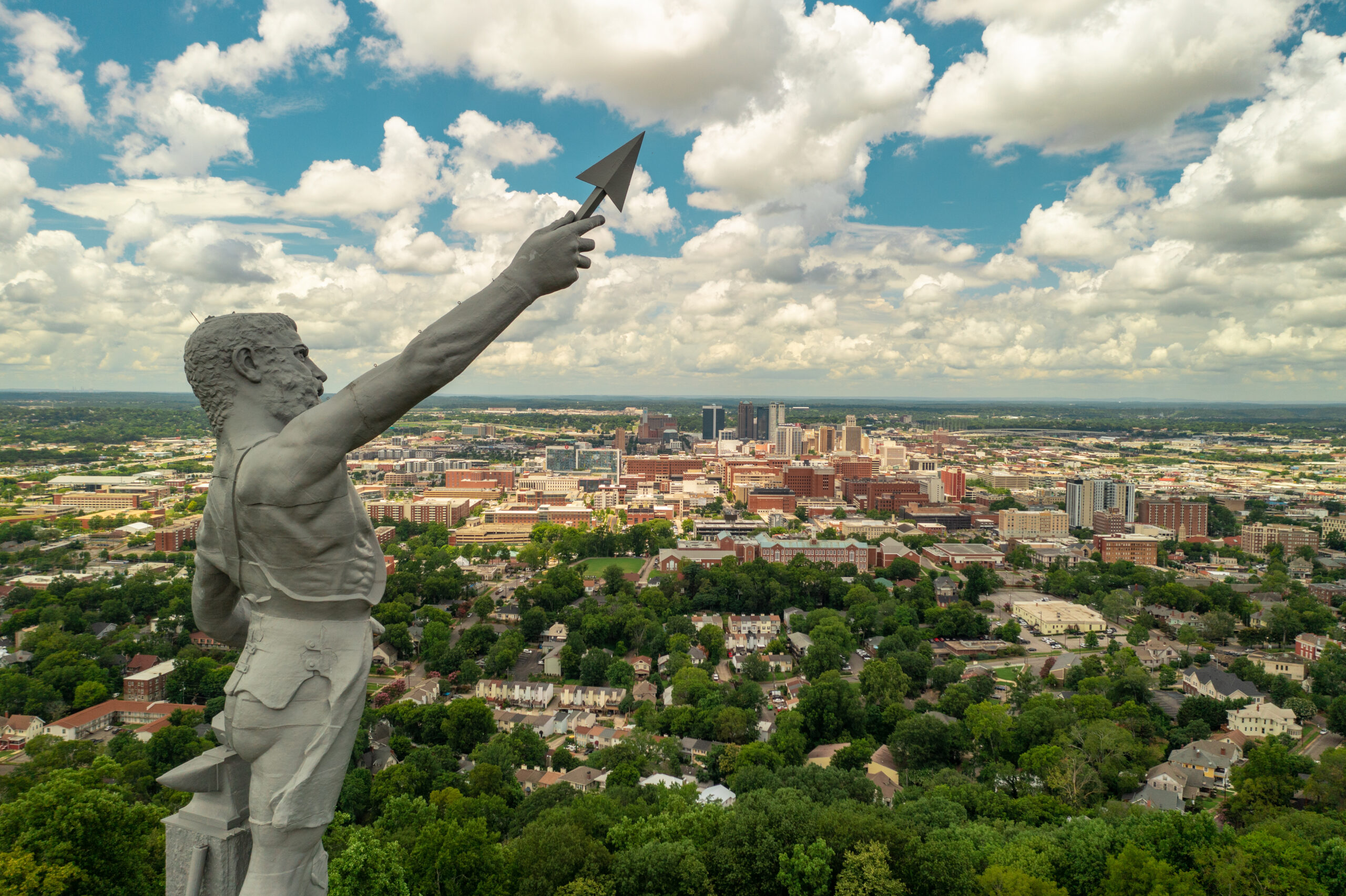 Aerial view of Birmingham, Alabama with the Vulcan statue prominently in the foreground, pointing towards the downtown skyline under a partly cloudy sky.