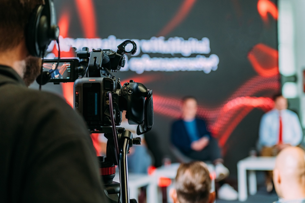 A camera operator recording a panel discussion, with the camera in focus and the panelists in the background out of focus against a red graphic backdrop.