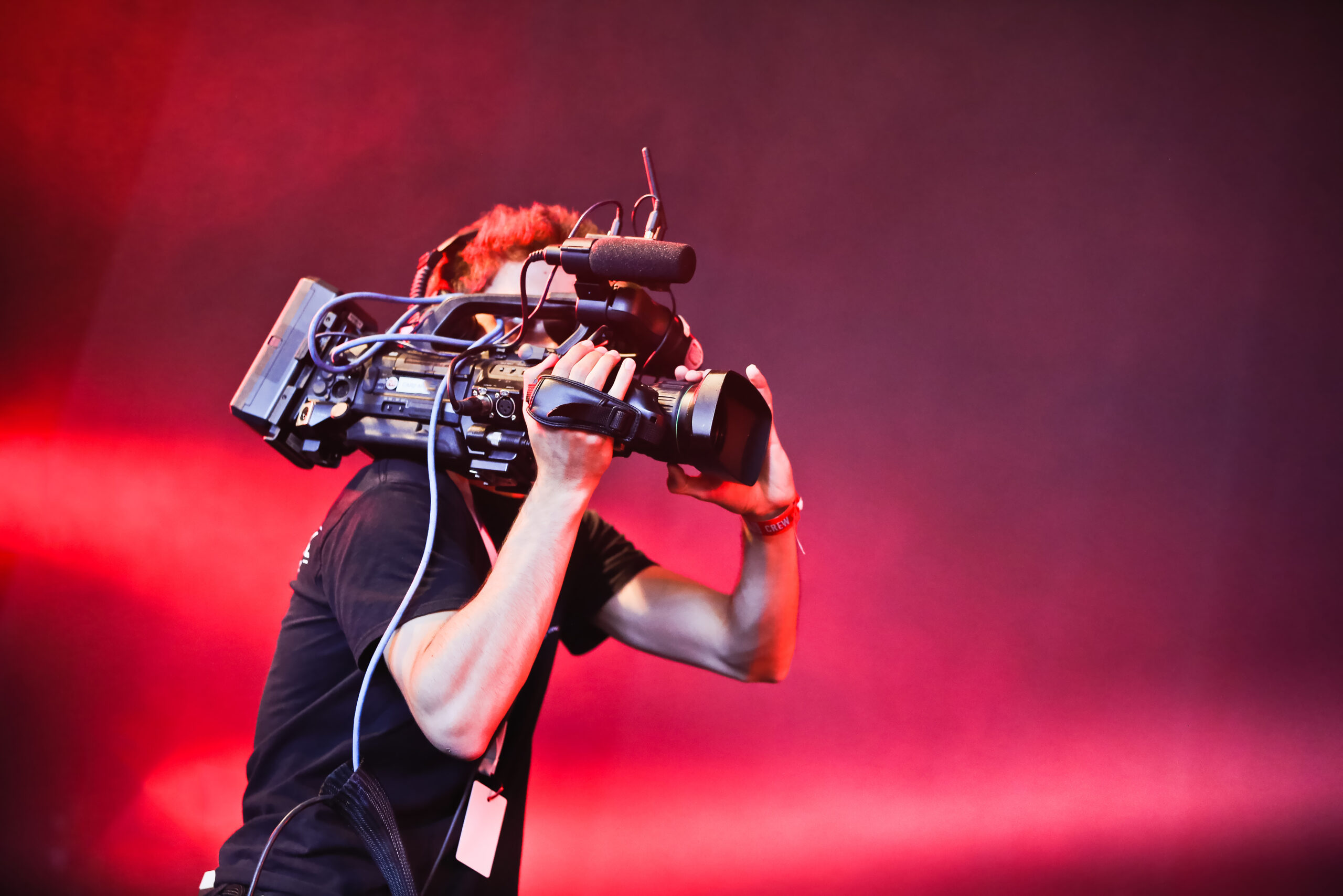 Professional camera operator filming on stage with a shoulder-mounted video camera, set against a red illuminated backdrop