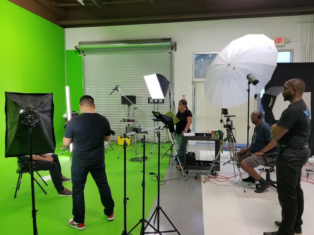 A busy production studio with a green screen setup. The scene includes various professional lighting equipment, cameras on tripods, a diverse crew of technicians and a director actively engaged in production tasks.