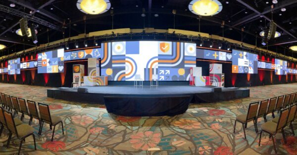 A wide conference stage with a high-resolution LED video wall displaying geometric blue, orange, and white graphics. The screen extends across the entire stage, with additional side panels showing matching visuals. Red stage lighting illuminates the black backdrop. Rows of chairs are set up for attendees, and truss-mounted speakers and lights are positioned above the stage.