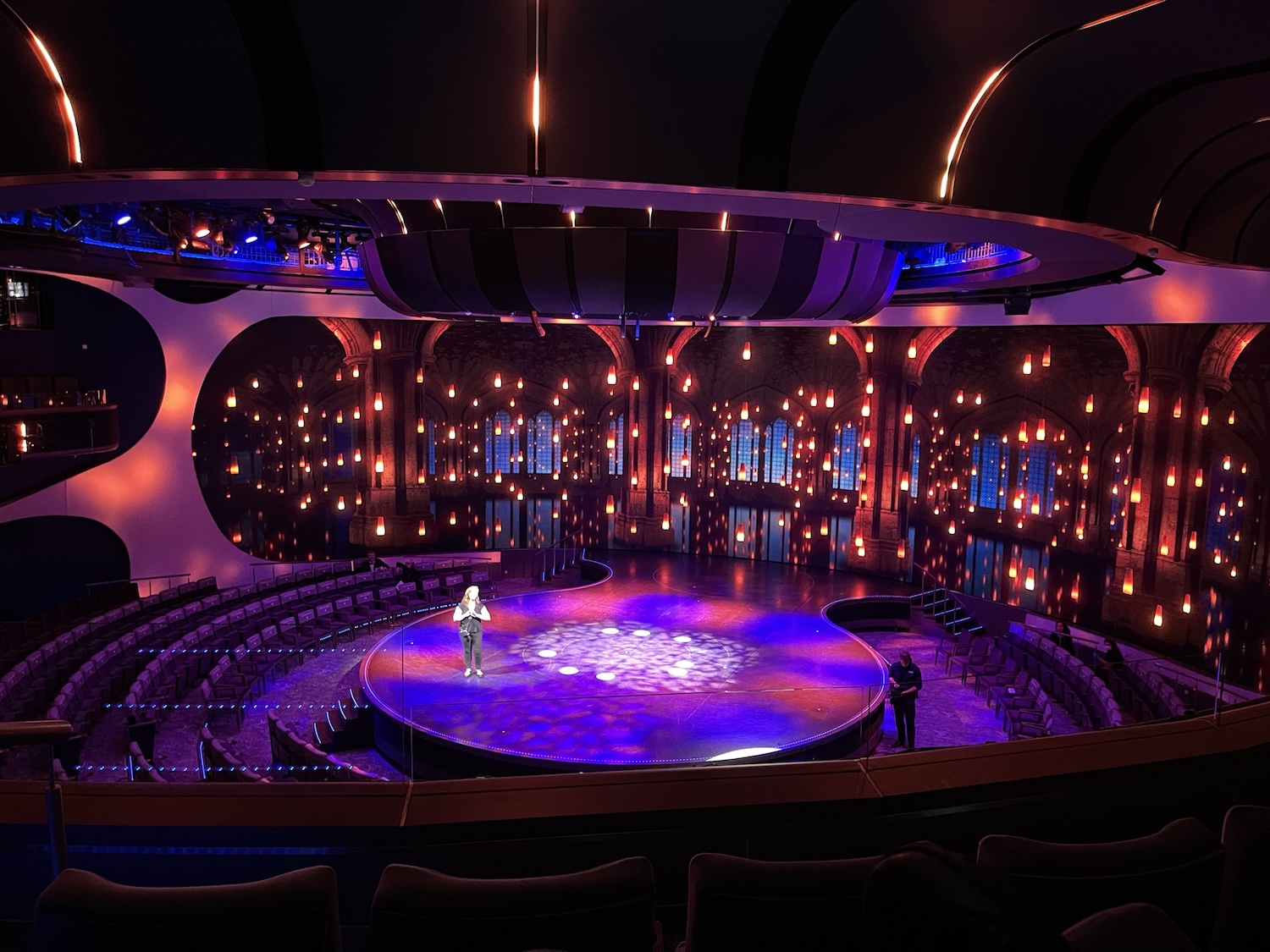 An opulent theater interior illuminated by blue and amber lights with a person standing on the central stage and another by the seating area.