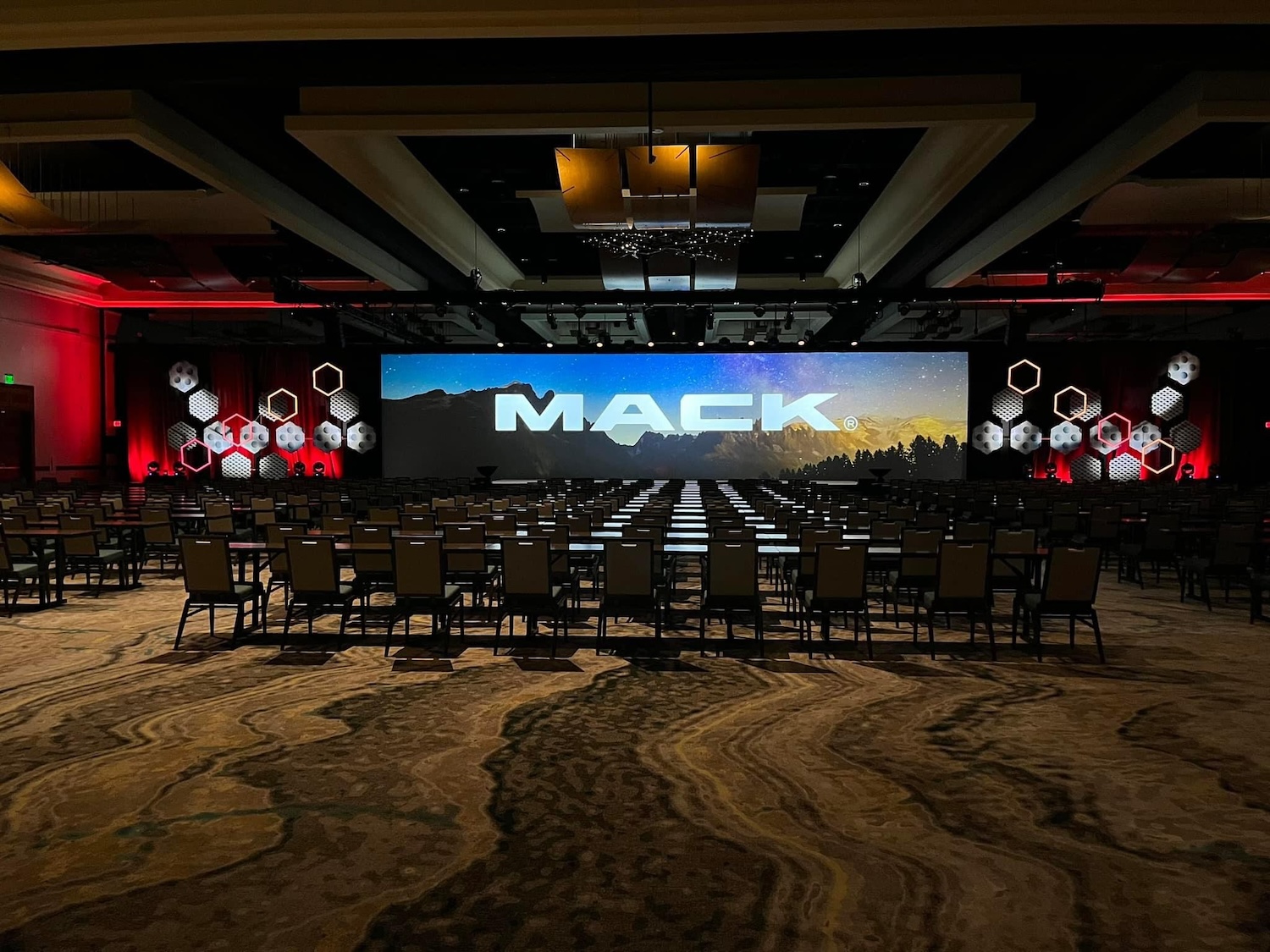 An event space with rows of chairs and a large screen displaying the 'MACK' logo, flanked by red lighting and geometric decor on the walls.