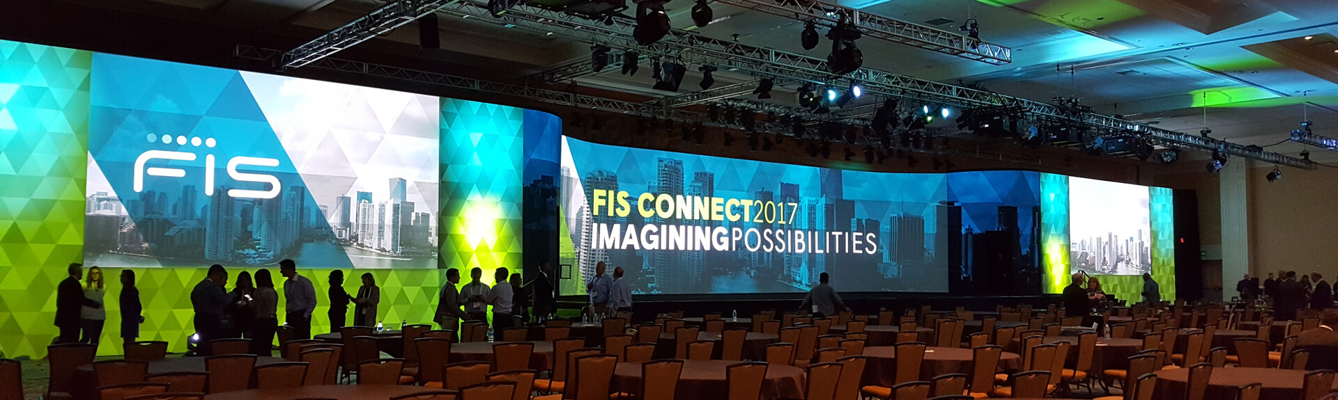 A large conference room set up for the FIS Connect 2017 event, featuring a massive curved LED screen displaying 'IMAGINING POSSIBILITIES' with a cityscape background. The screen is framed by geometric green and blue lighting. Several people are standing near the stage, while tables and chairs fill the foreground. Overhead trusses support stage lighting and AV equipment.