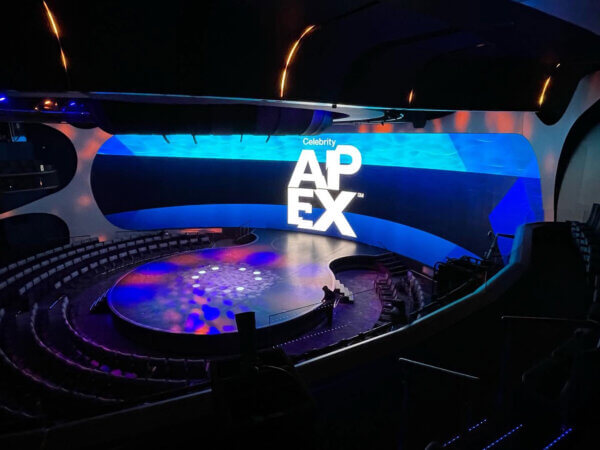 A modern theater stage with a curved LED video wall displaying 'Celebrity APEX' in bold white letters on a blue background. The circular stage is illuminated with blue and purple lighting effects, and the seating area is arranged in a semi-circle around it. The ceiling features sleek, curved architectural elements with integrated lighting. A person is visible near the stage, likely making final adjustments to the setup. The AV design includes an expansive high-resolution screen and immersive lighting.