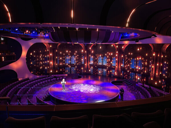 theater stage with a circular design, illuminated by blue and purple stage lighting. A presenter stands in the center, addressing an empty auditorium with curved seating. The backdrop features an LED screen displaying an ornate, cathedral-like scene with hanging lanterns and arched windows. The ceiling includes modern architectural elements with integrated lighting.