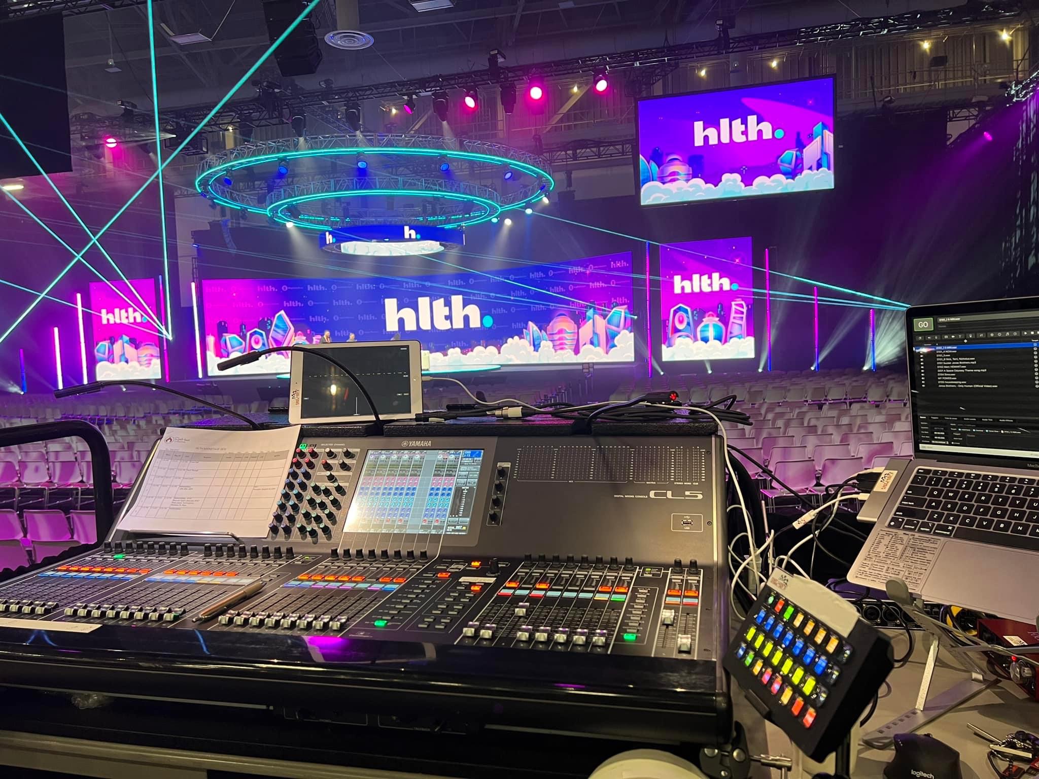 Control panel of an event production setup with sound and light mixing boards, overlooking an empty venue with chairs and large screens displaying the text "hlth."