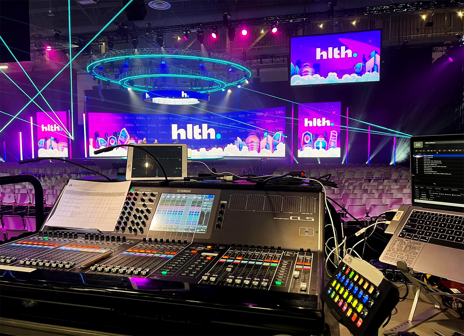 A sound and lighting control desk with a view of a stage and screens displaying 'hlth' branding, bathed in vibrant stage lighting.