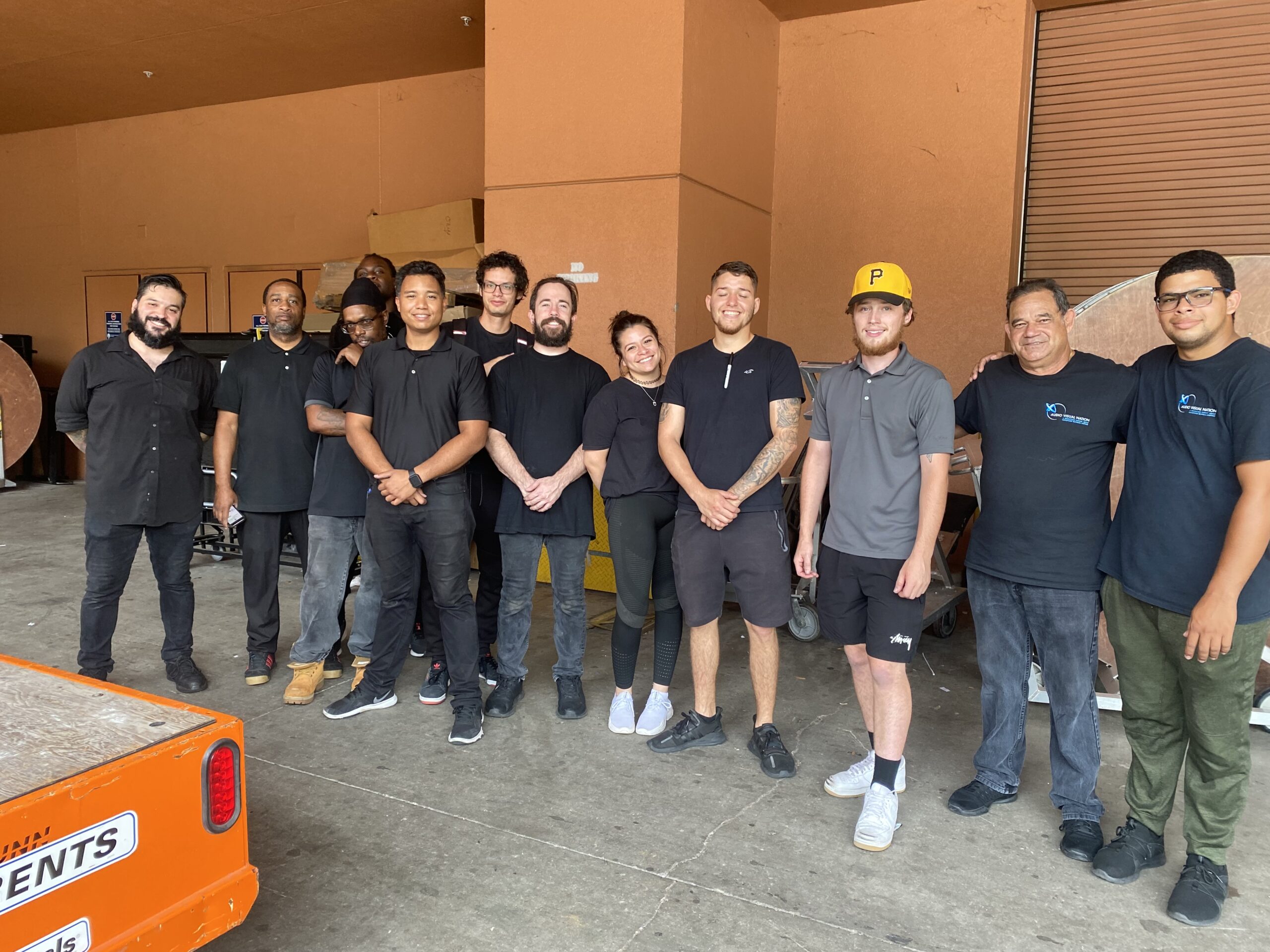 A group photo of Audio Visual Nation crew members, all dressed in black, smiling and standing together outside a loading dock.
