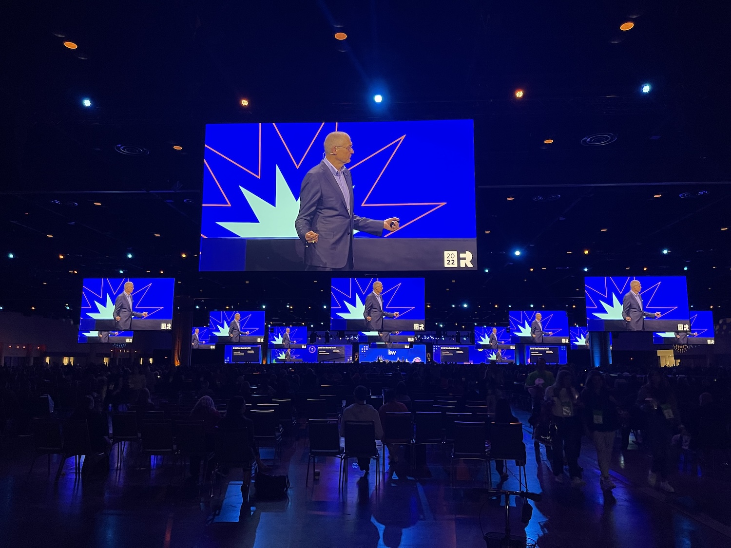 A conference room with a speaker on stage, displayed on multiple large screens with a blue and white graphic design, and an audience in a large event space.