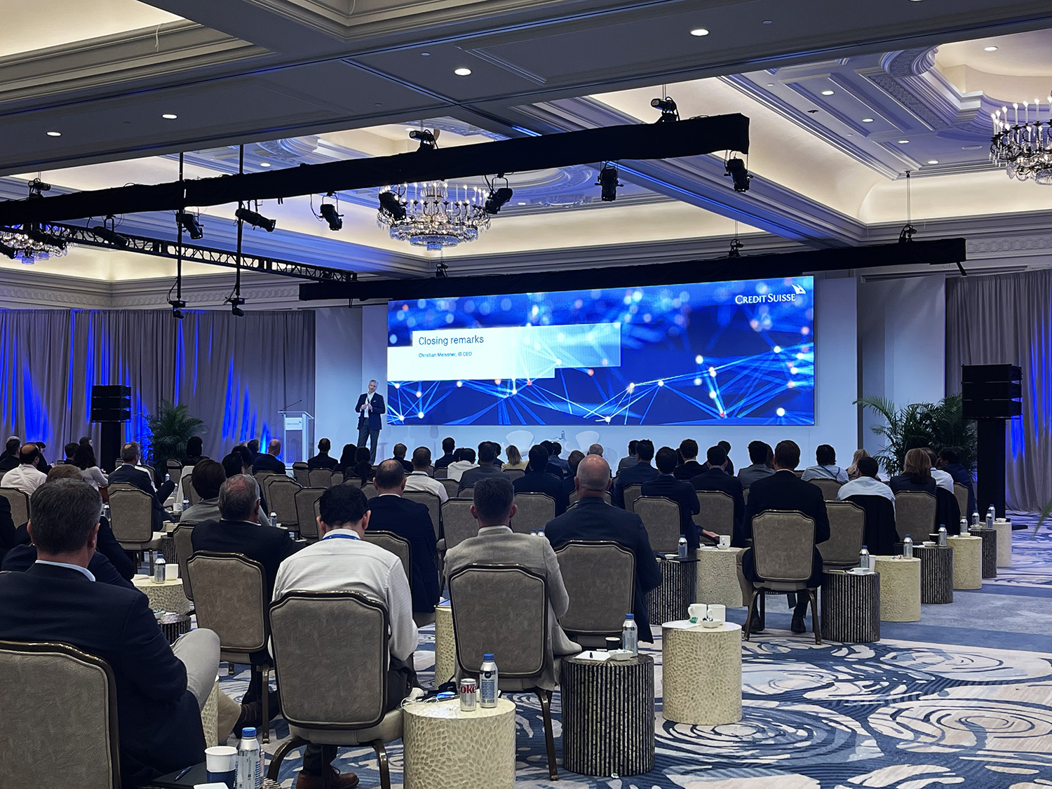 An elegant conference room with attendees facing a stage where a speaker is presenting, with a large screen displaying 'Closing remarks' and a 'Credit Suisse' background.