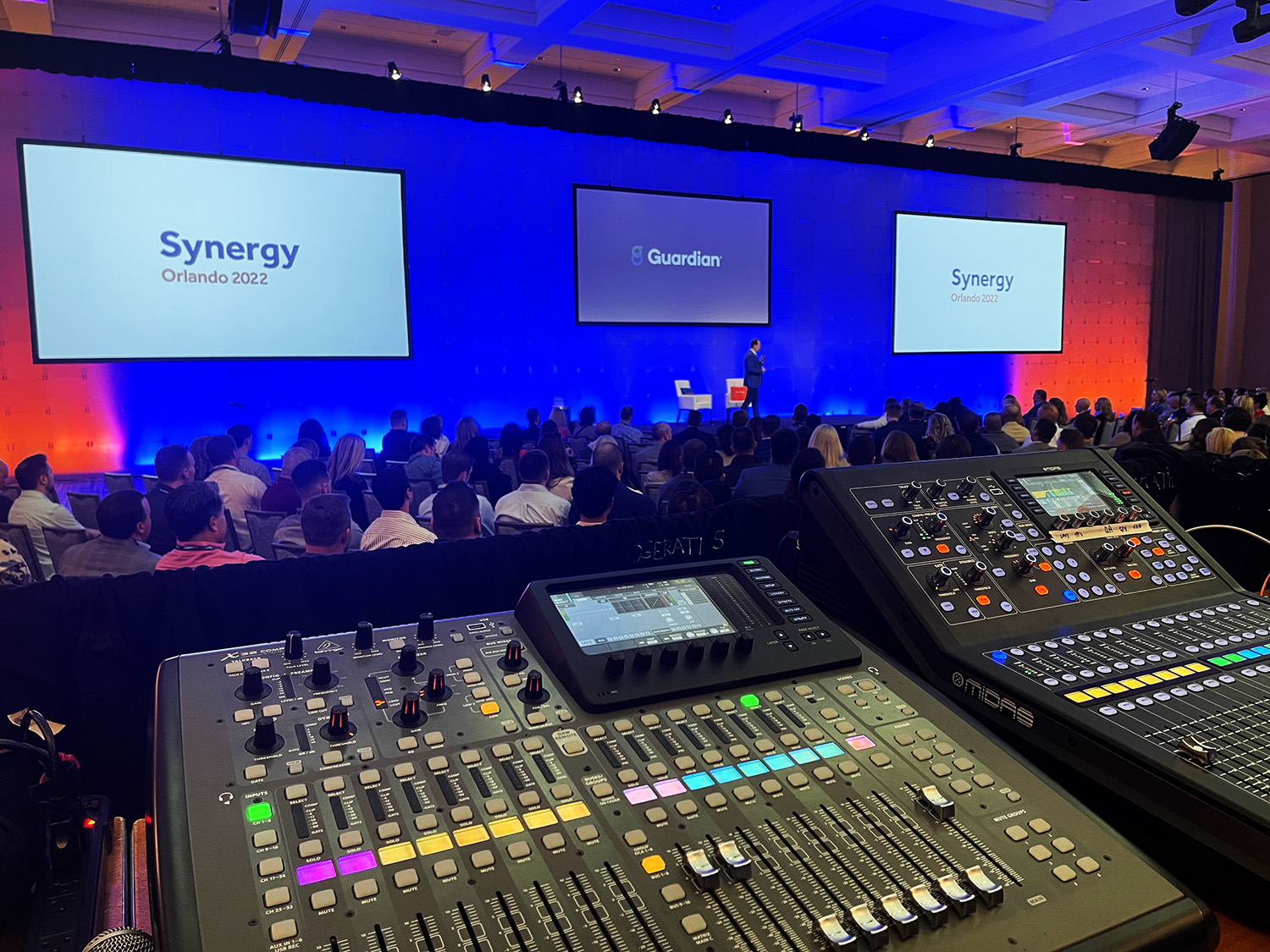 A sound mixing console in the foreground with an audience and presenter at the 'Synergy Orlando 2022' conference in the background.