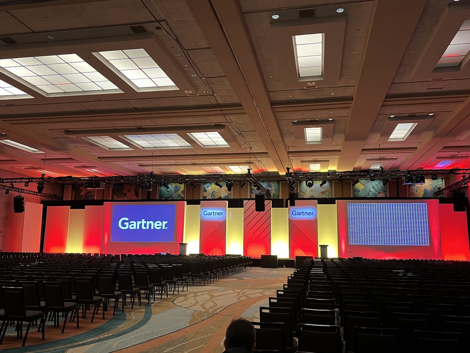 A spacious conference room with rows of chairs facing a stage, lit with red tones and featuring screens with 'Gartner' logos.
