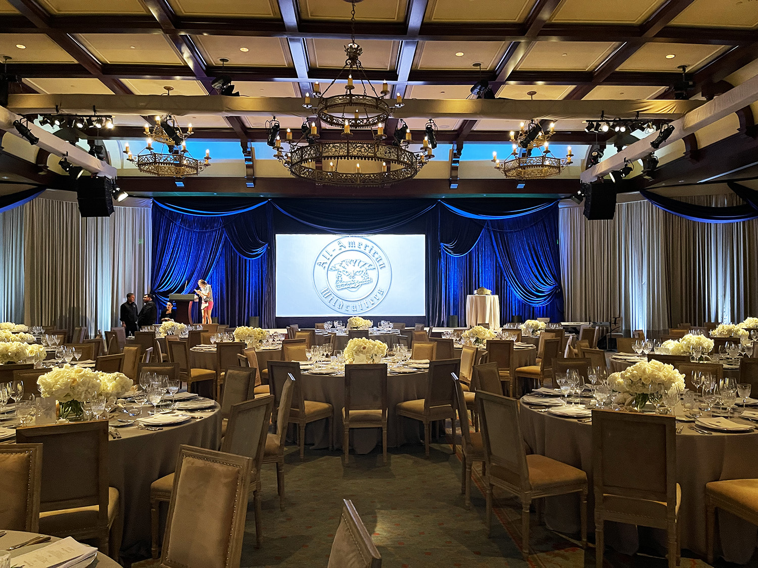 An elegant banquet hall set up for an event with round tables, floral centerpieces, a stage with a blue backdrop, and a large screen displaying an emblem.