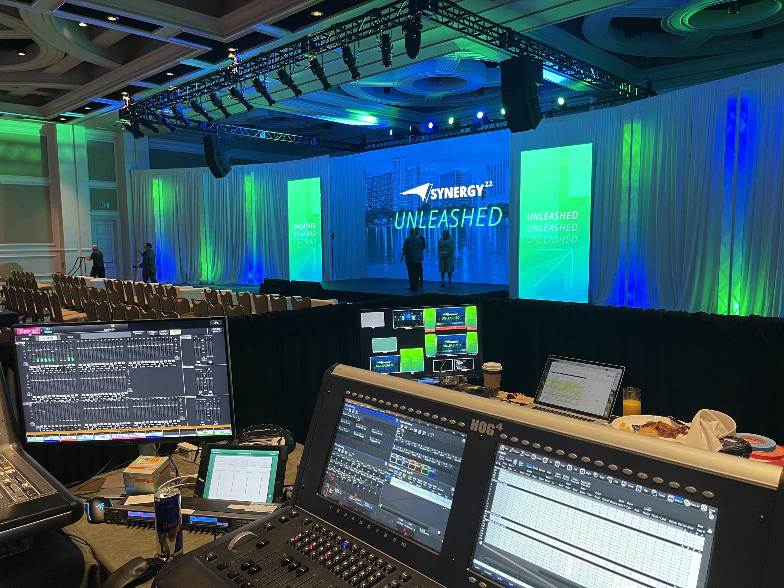 A control booth with multiple monitors and lighting consoles overlooks a large conference stage. The stage features the Synergy logo and the word "UNLEASHED" on a bright screen, with vibrant green and blue lighting illuminating the curtains in the background.