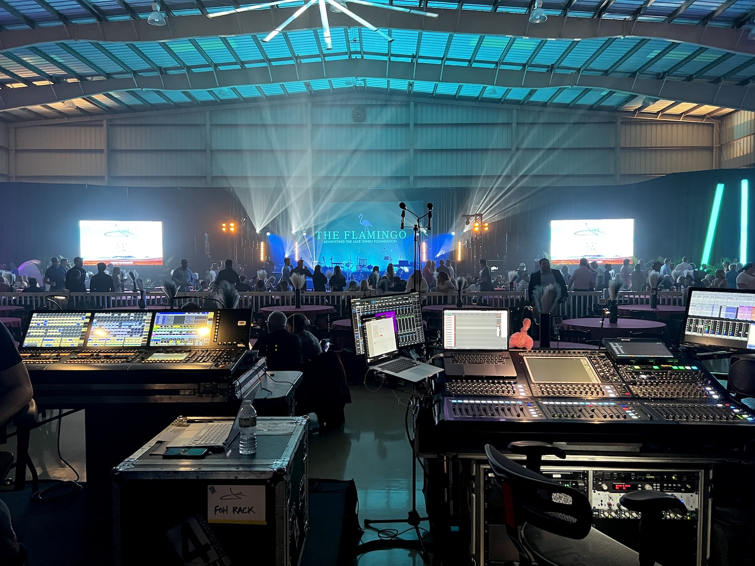 A technical production setup with multiple control boards in the foreground, overlooking an event space with the stage lit up and the text 'THE FLAMINGO' prominently displayed.