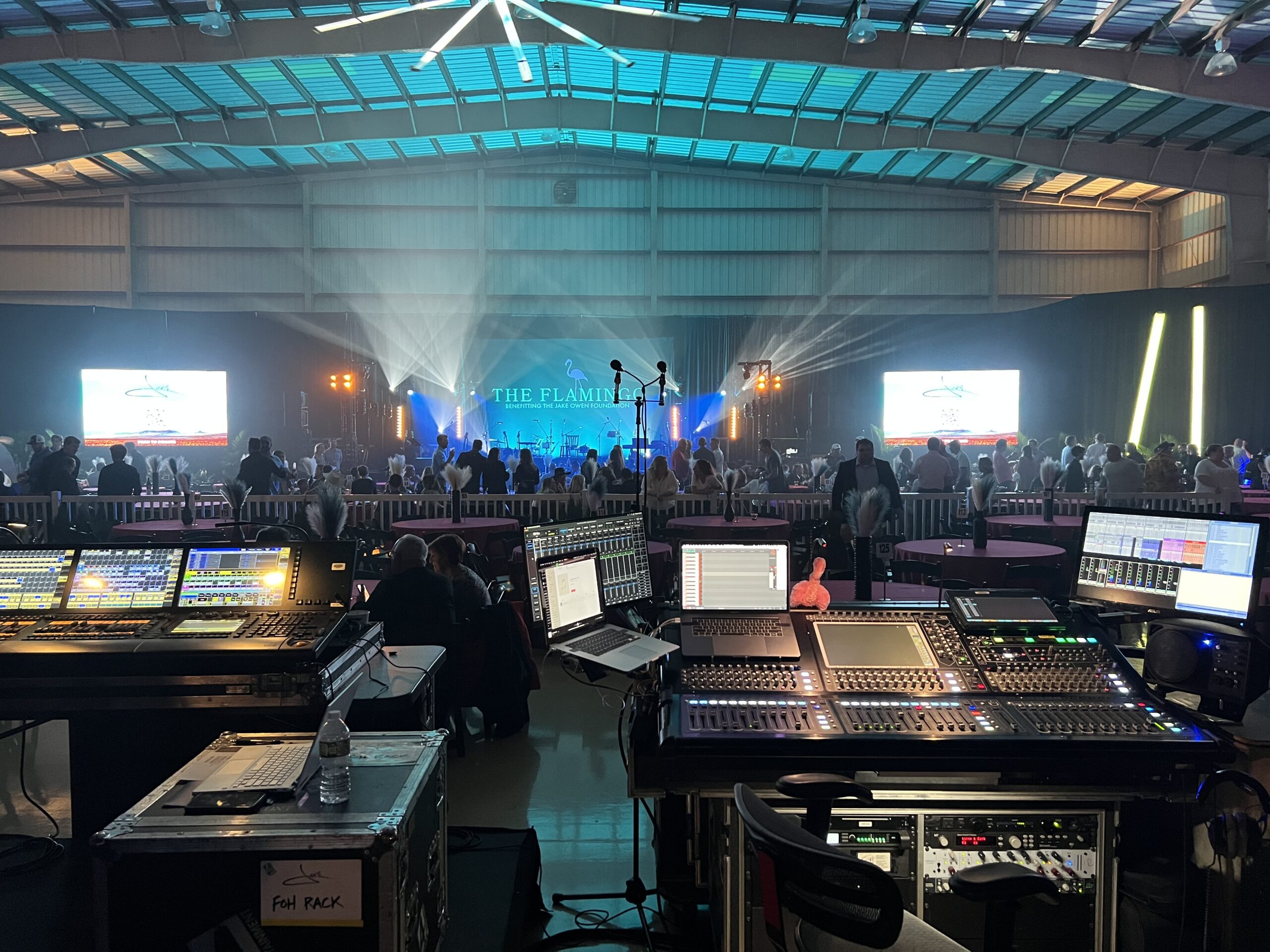 An indoor concert event featuring multiple mixing consoles and digital audio workstations in the foreground, with a crowded audience and a live performance in the background illuminated by stage lights.