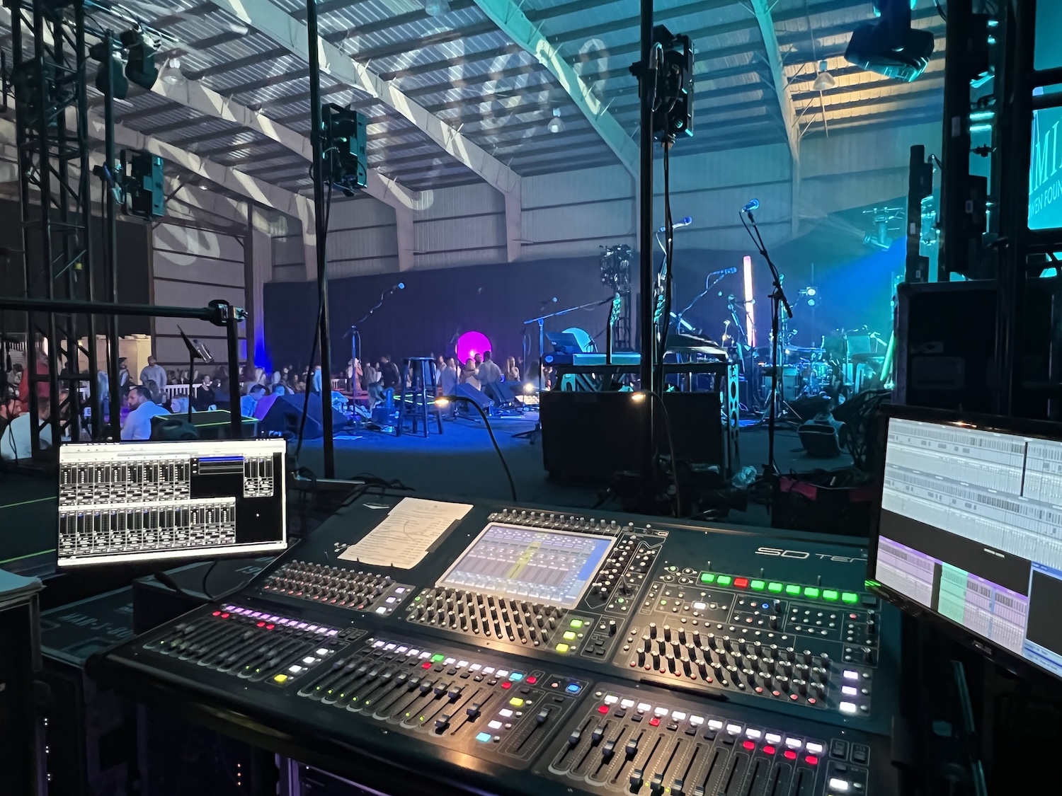 A view over a sound mixing console at a live performance space with musical instruments set up on stage and an audience in the background.