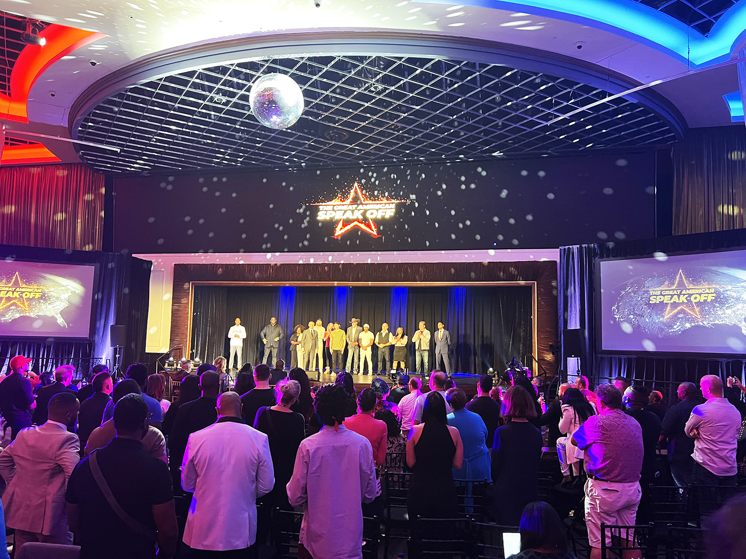 A vibrant event with a standing audience applauding a group on stage, under a disco ball, with screens displaying 'SPEAK OFF' and starry lighting effects.