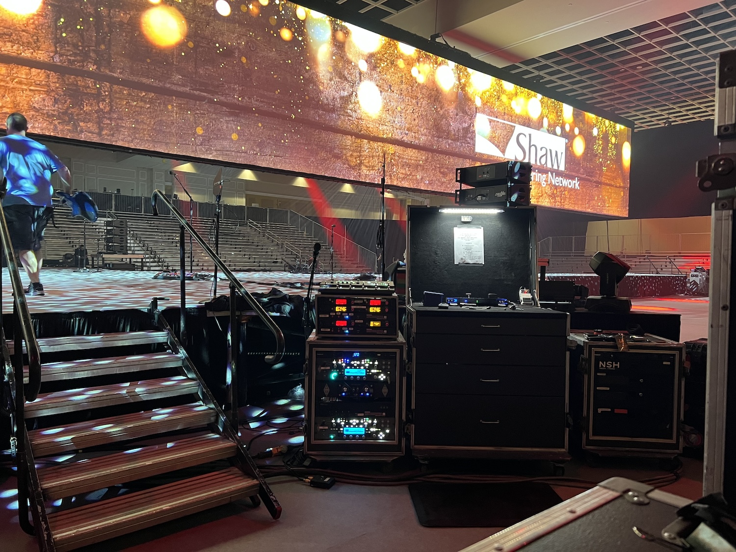 Backstage view at an event with technical equipment and racks, a large screen with 'Shaw Connecting Network' branding, and steps leading up to an empty stage with seating in the background.