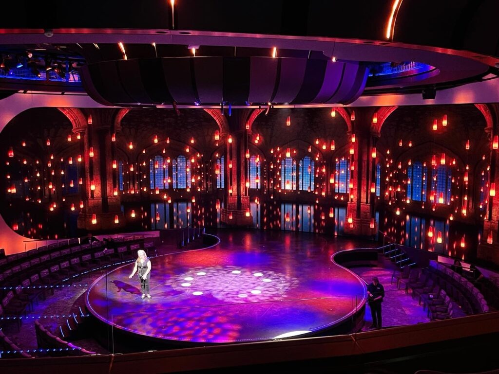 A theater with a circular stage bathed in blue and red lights, surrounded by tiered seating, and a person standing in the spotlight.