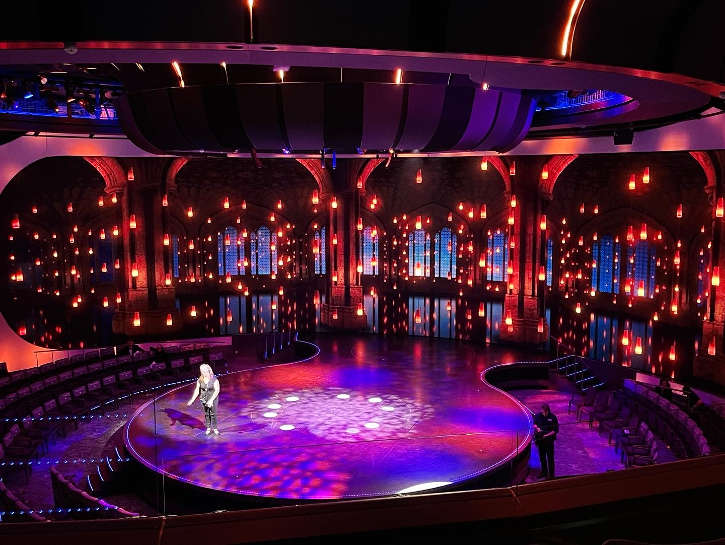 A theater with a circular stage bathed in blue and red lights, surrounded by tiered seating, and a person standing in the spotlight.