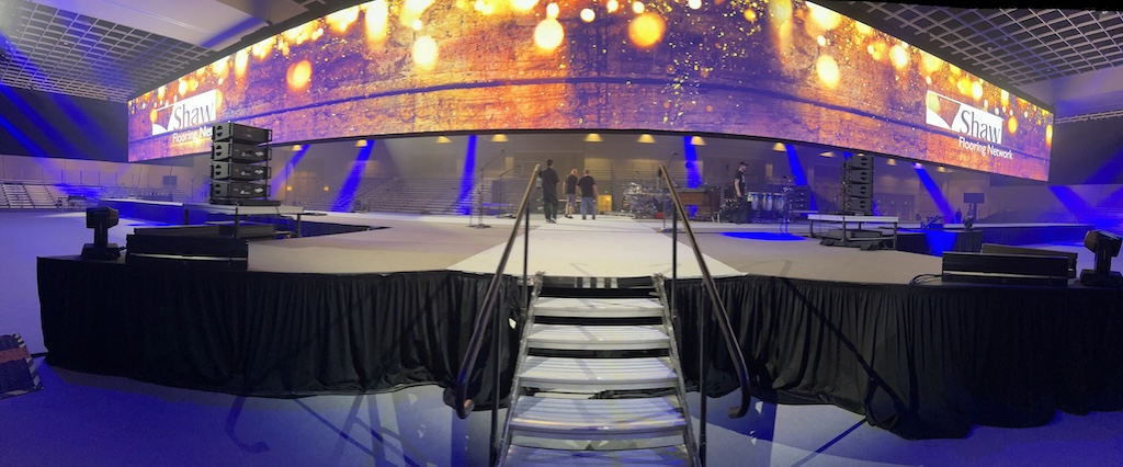 A wide stage setup for 'Shaw Flooring Network' featuring a massive, curved digital display showing a vibrant, textured visual. The stage is equipped with professional lighting and sound equipment, prepared for an event with seating in the background.