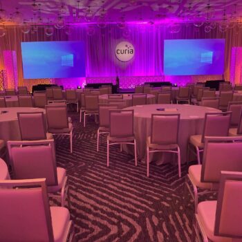 A conference room with round tables and chairs is set up facing two large projection screens displaying a Windows desktop. The room is illuminated with purple and orange lighting, and a "Curia" logo is displayed on a circular sign above the stage.